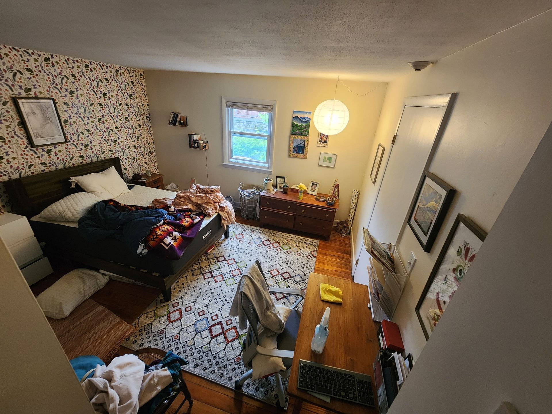 Bedroom with unmade bed, patterned wallpaper, wooden desk and dresser, rug, and window.