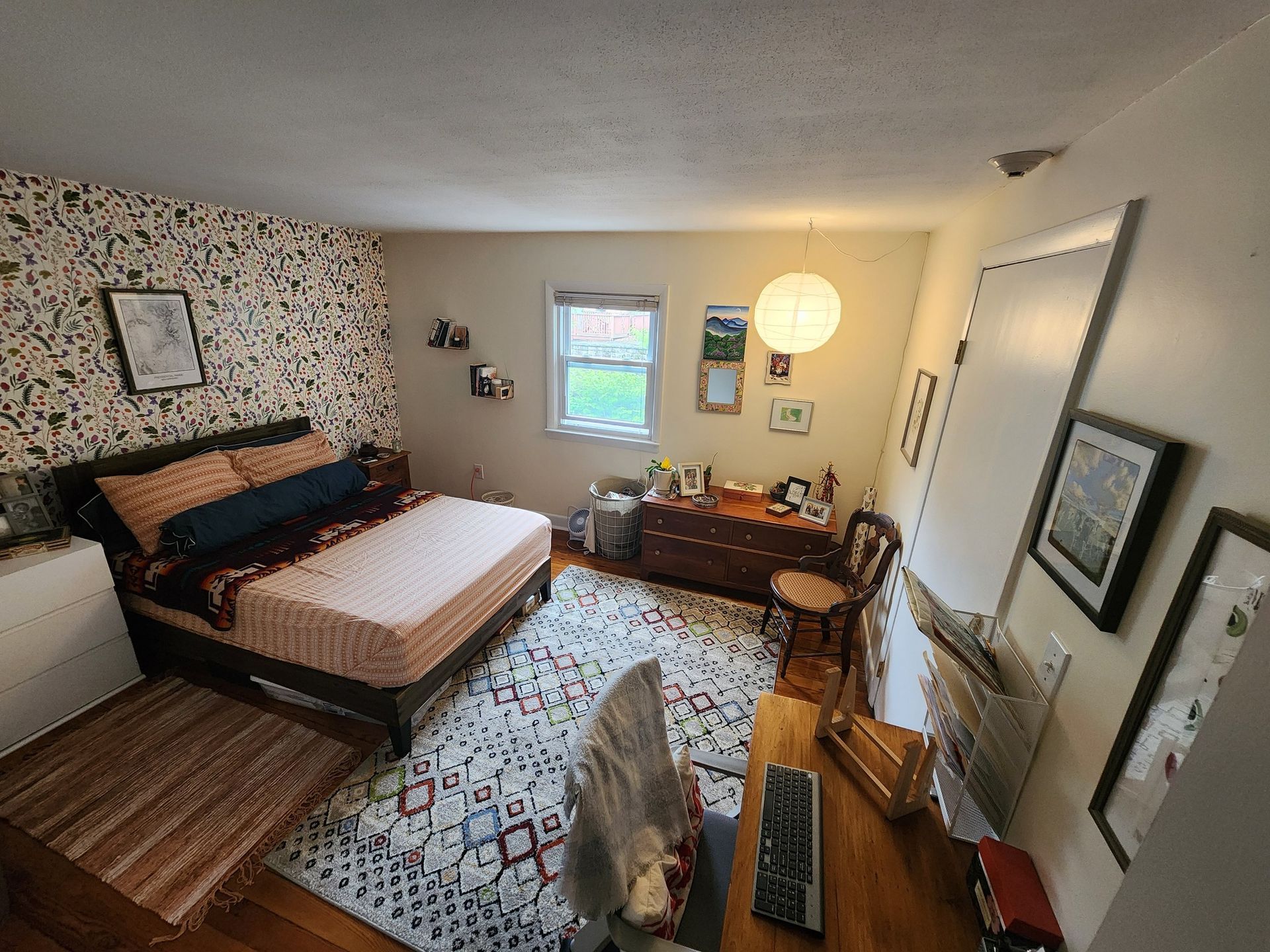 Bedroom with bed, desk, rug, and floral wallpaper. Light fixture and window present.