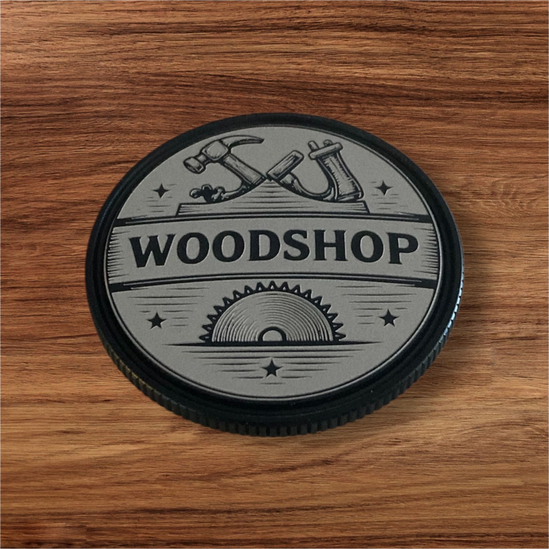 Alt text:
Round engraved woodshop coin featuring woodworking tools and a saw blade design with the word “WOODSHOP,” set on a wood surface.
