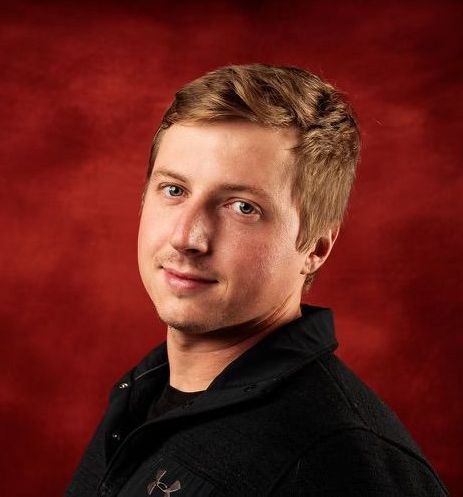 A young man in a black shirt is standing in front of a red background.