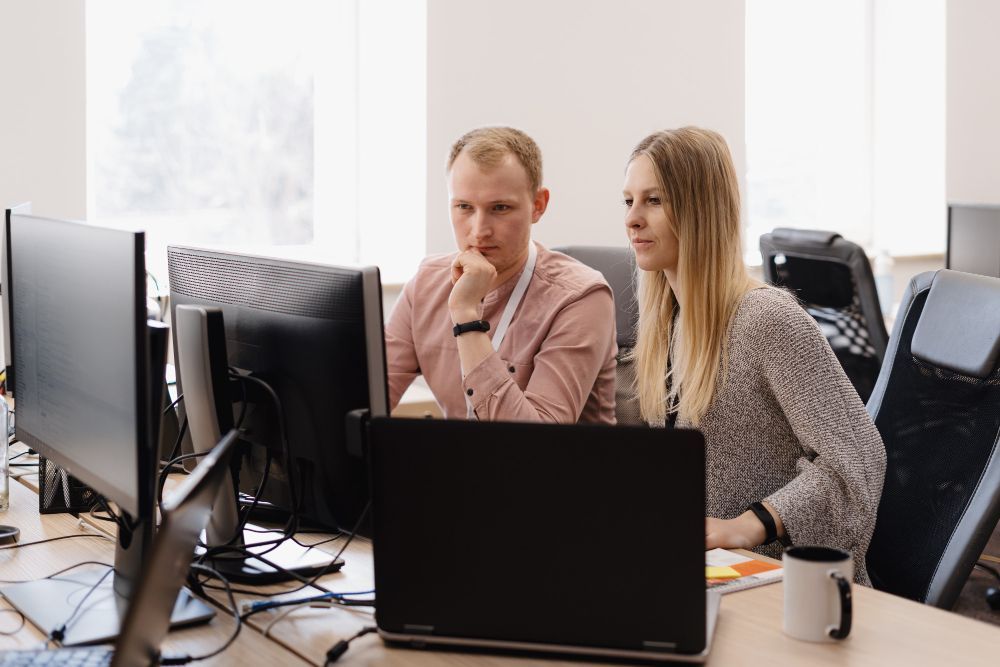 A man and a woman are sitting at a desk looking at a computer screen.