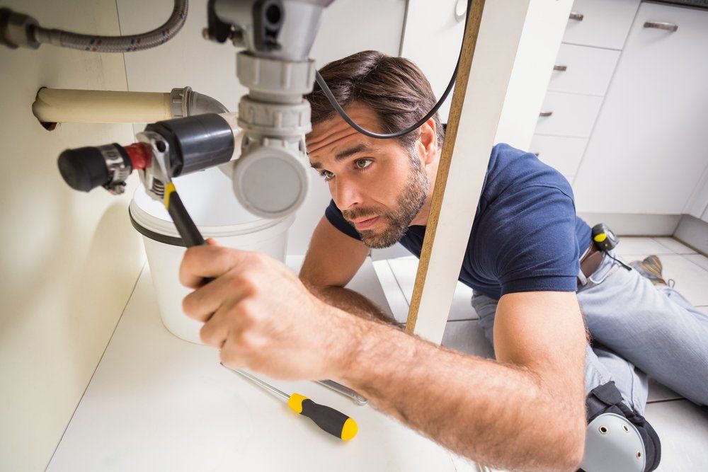 A Man is Kneeling on the Floor Fixing a Sink Pipe  — SOS Hot Water & Plumbing Services in Kiama Downs, NSW