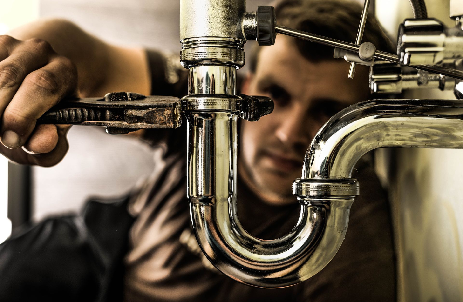 Closeup of emergency plumber using a wrench to tighten a metallic siphon under a sink. Closeup of emergency plumber using a wrench to tighten a metallic siphon under a sink.