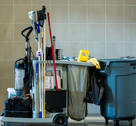 Un carrito de limpieza gris, repleto de fregonas, una aspiradora, suministros y un gran cubo de basura, está apoyado contra una pared de azulejos.