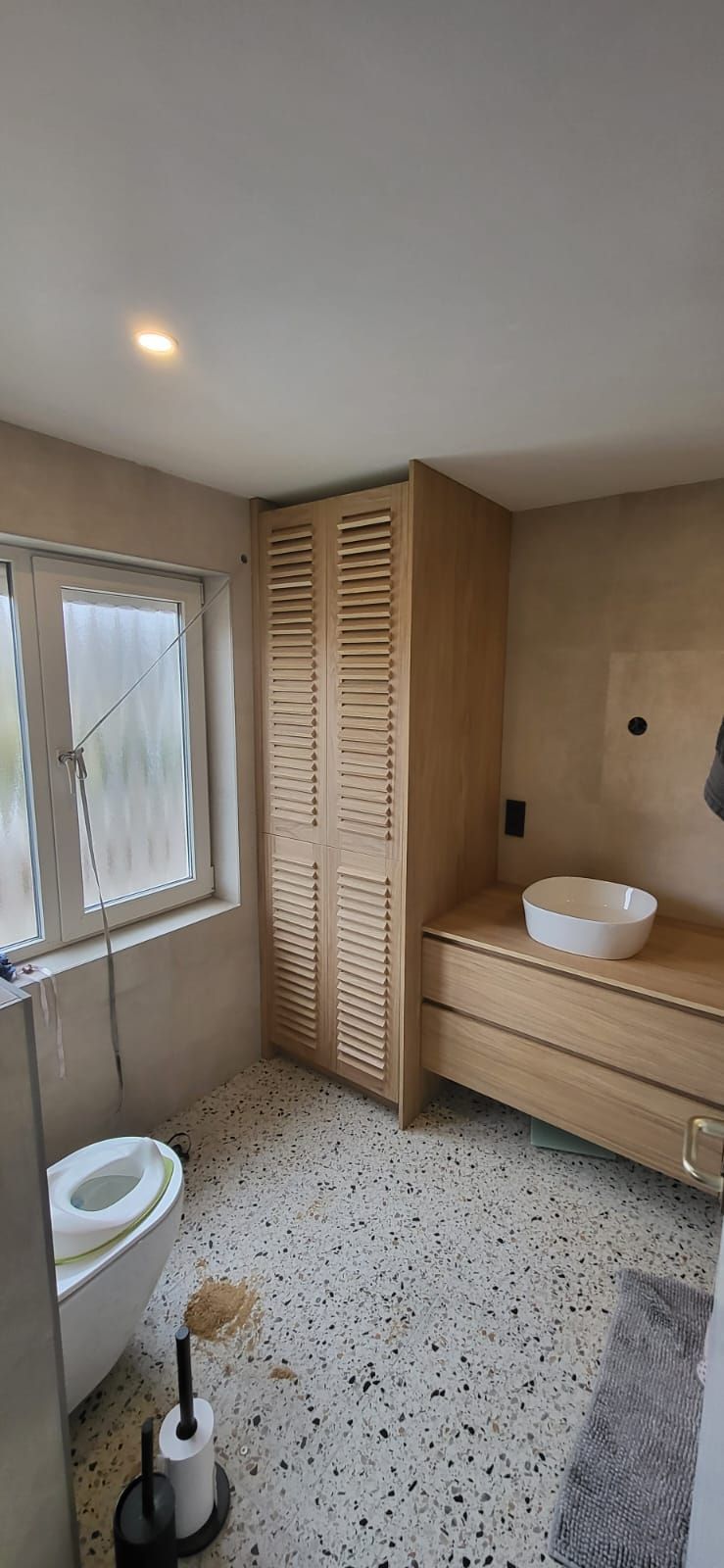 Een badkamer met houten kasten, een wastafel en een toilet. De vloer heeft een gespikkeld patroon.