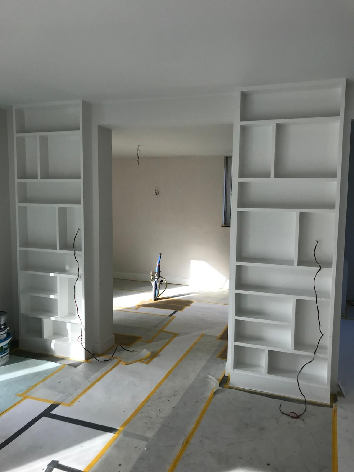 Witte ingebouwde boekenplanken omlijsten een deuropening in een kamer in aanbouw. ​​Er hangen draden en de vloer is afgeplakt.
