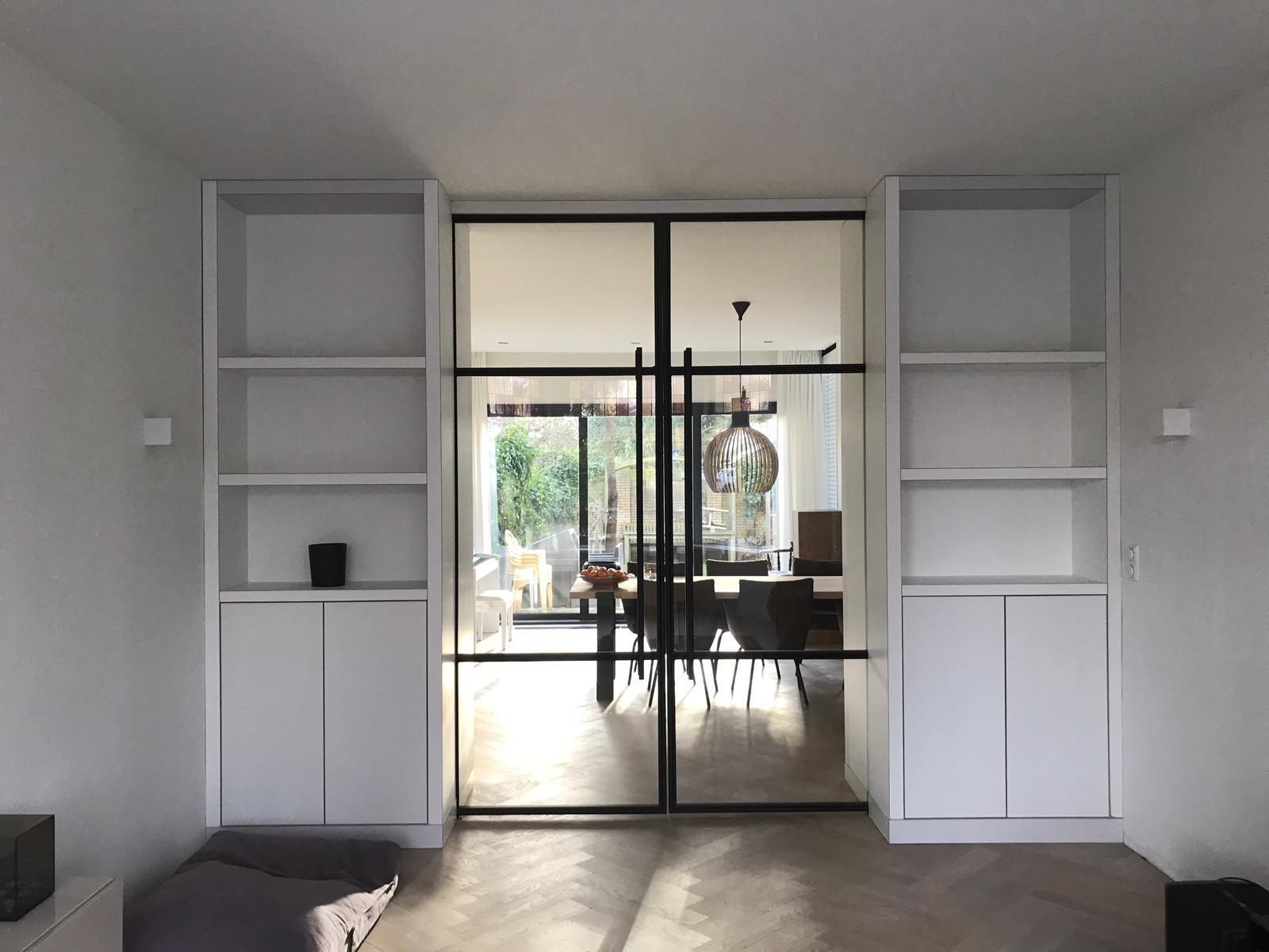 Ingebouwde witte planken aan weerszijden van een zwarte glazen deur, die toegang geeft tot een eethoek met een tafel en stoelen.