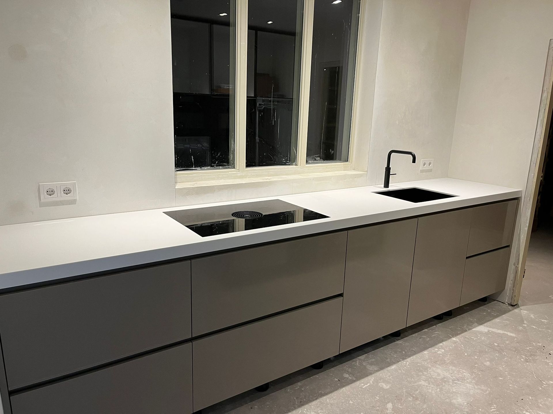 Moderne keuken met grijze kasten, wit aanrechtblad, zwarte spoelbak en kraan, raam.