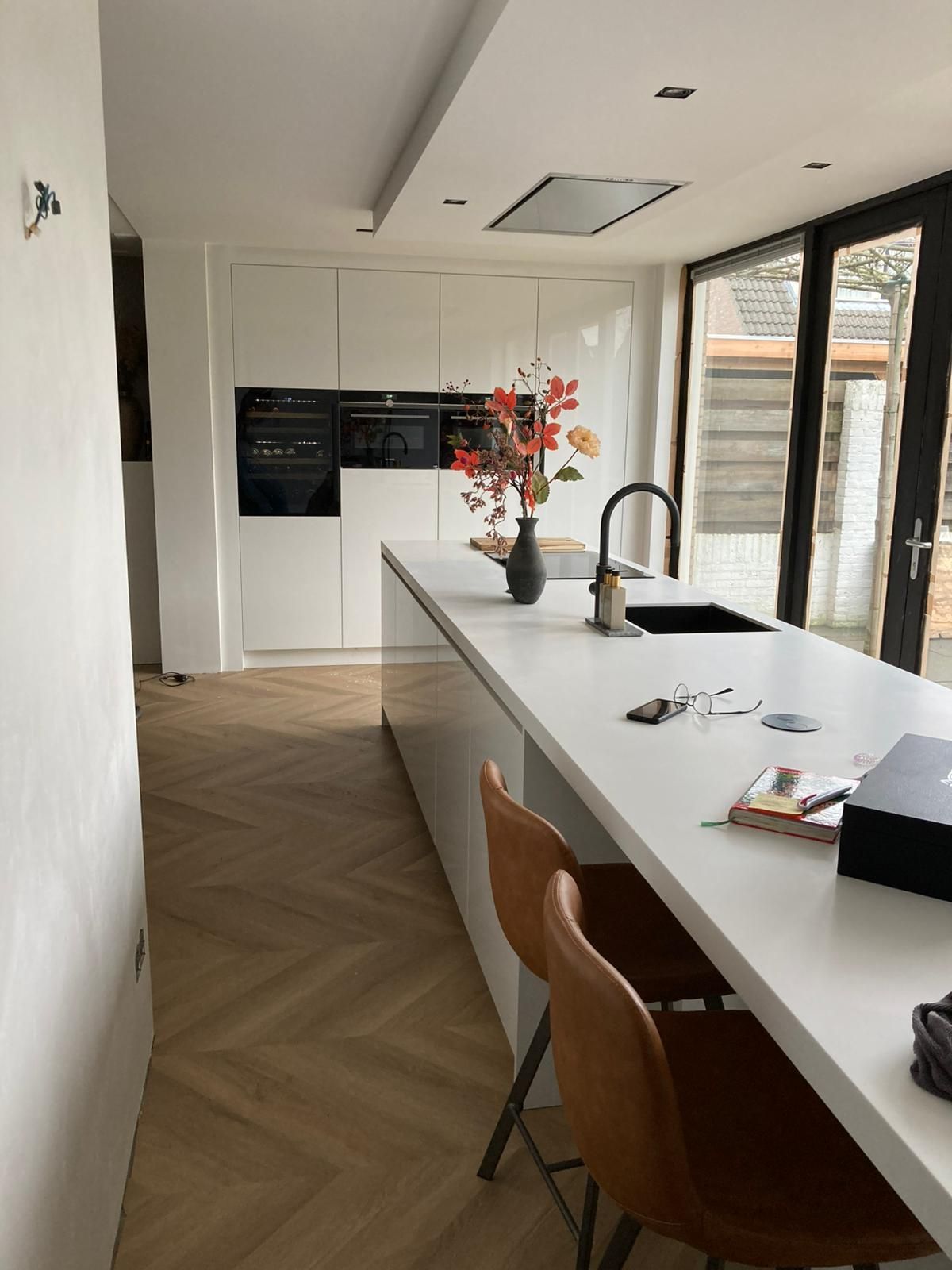 Moderne keuken met witte kasten en kookeiland, visgraatvloer en leren barkrukken.