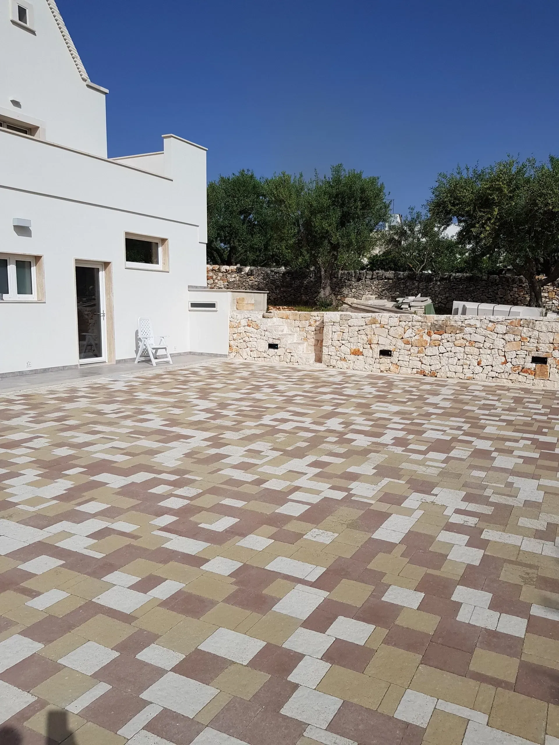 Edificio bianco con patio in pietra, mattoni rossi/beige. Alberi sullo sfondo, cielo azzurro.