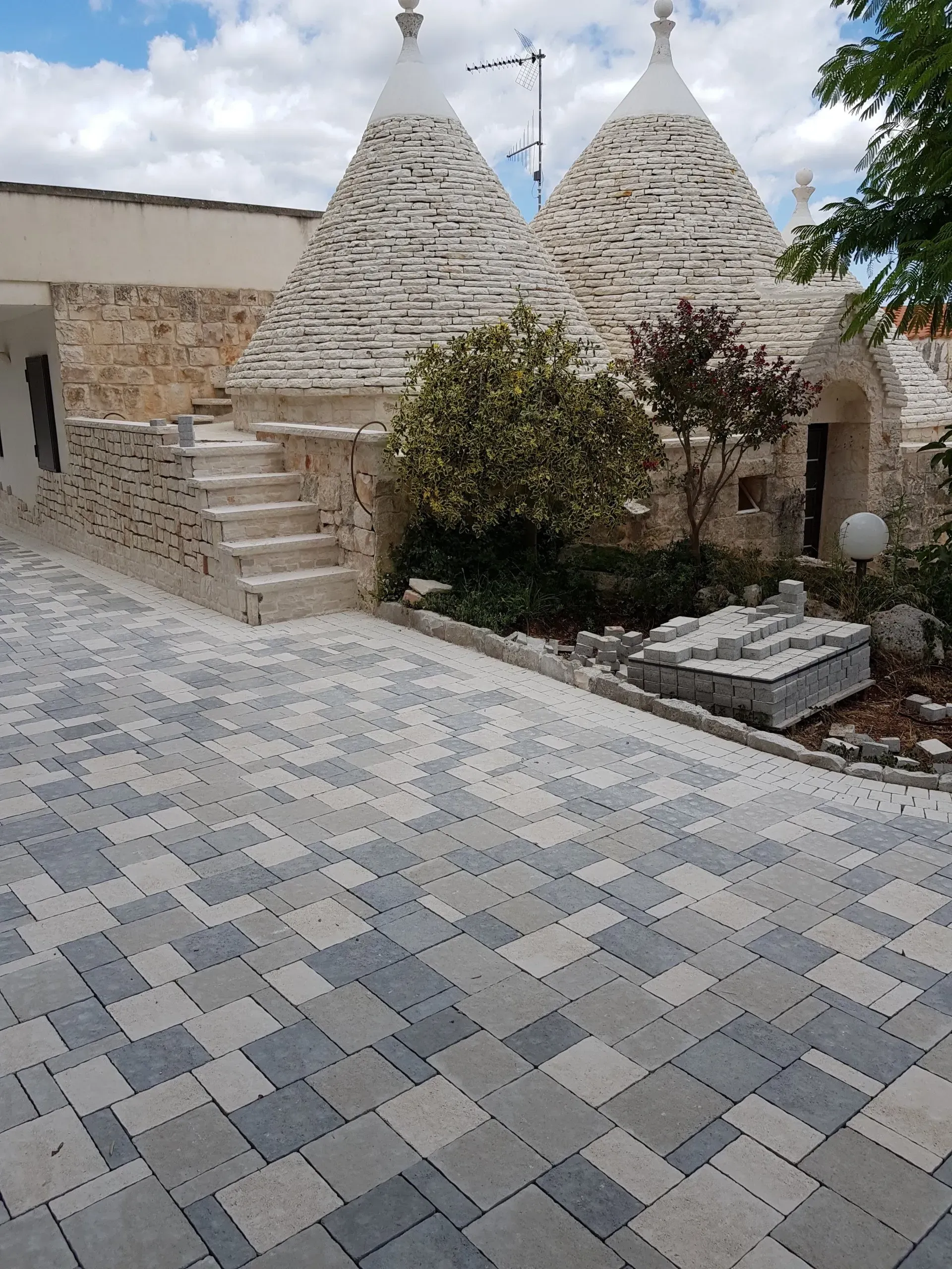 Trulli in pietra con tetti conici, vialetto in ciottoli bianchi e grigi, piccolo giardino.