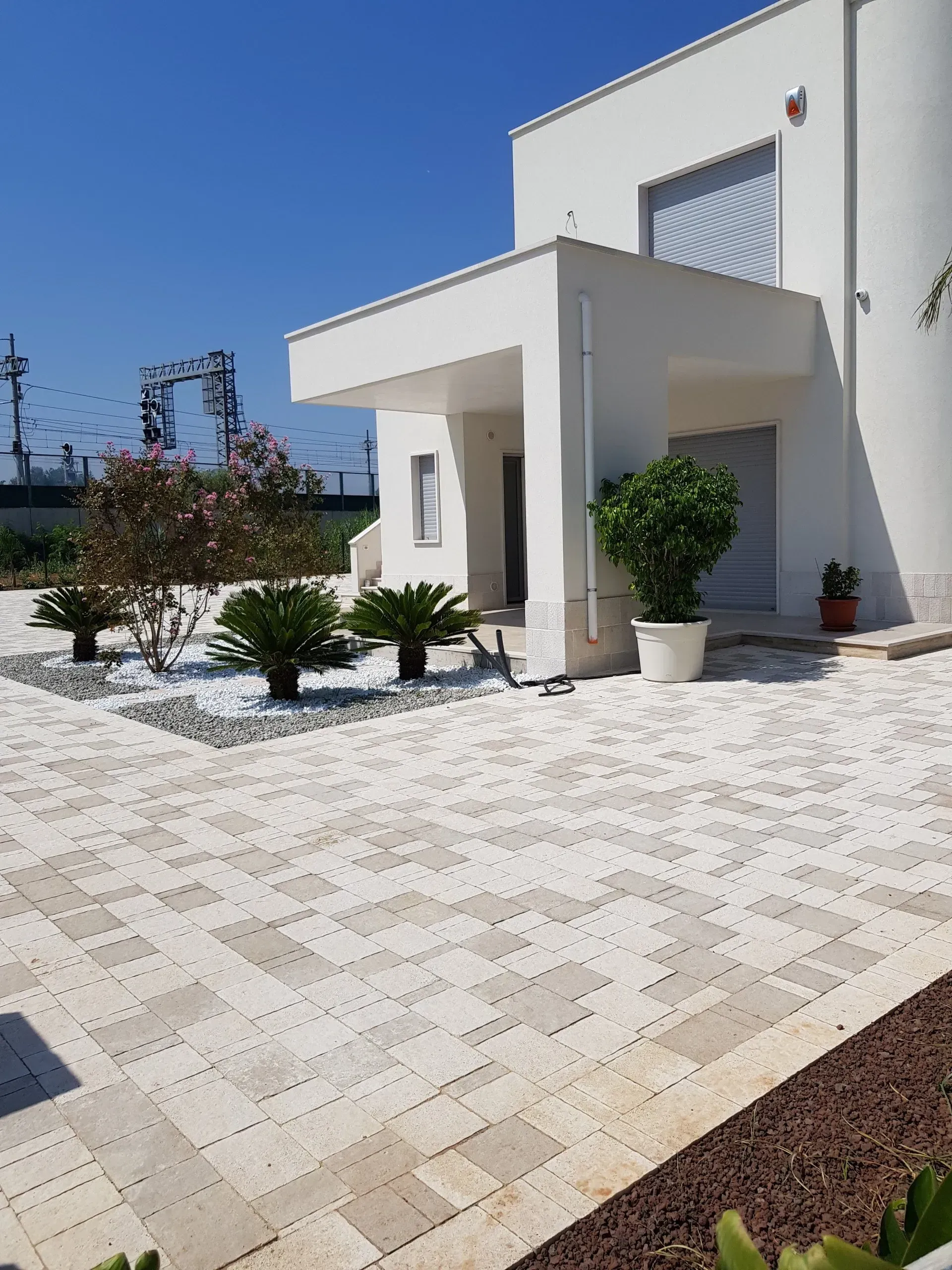 Moderna casa bianca con giardino pavimentato, piccoli alberi e cielo azzurro.