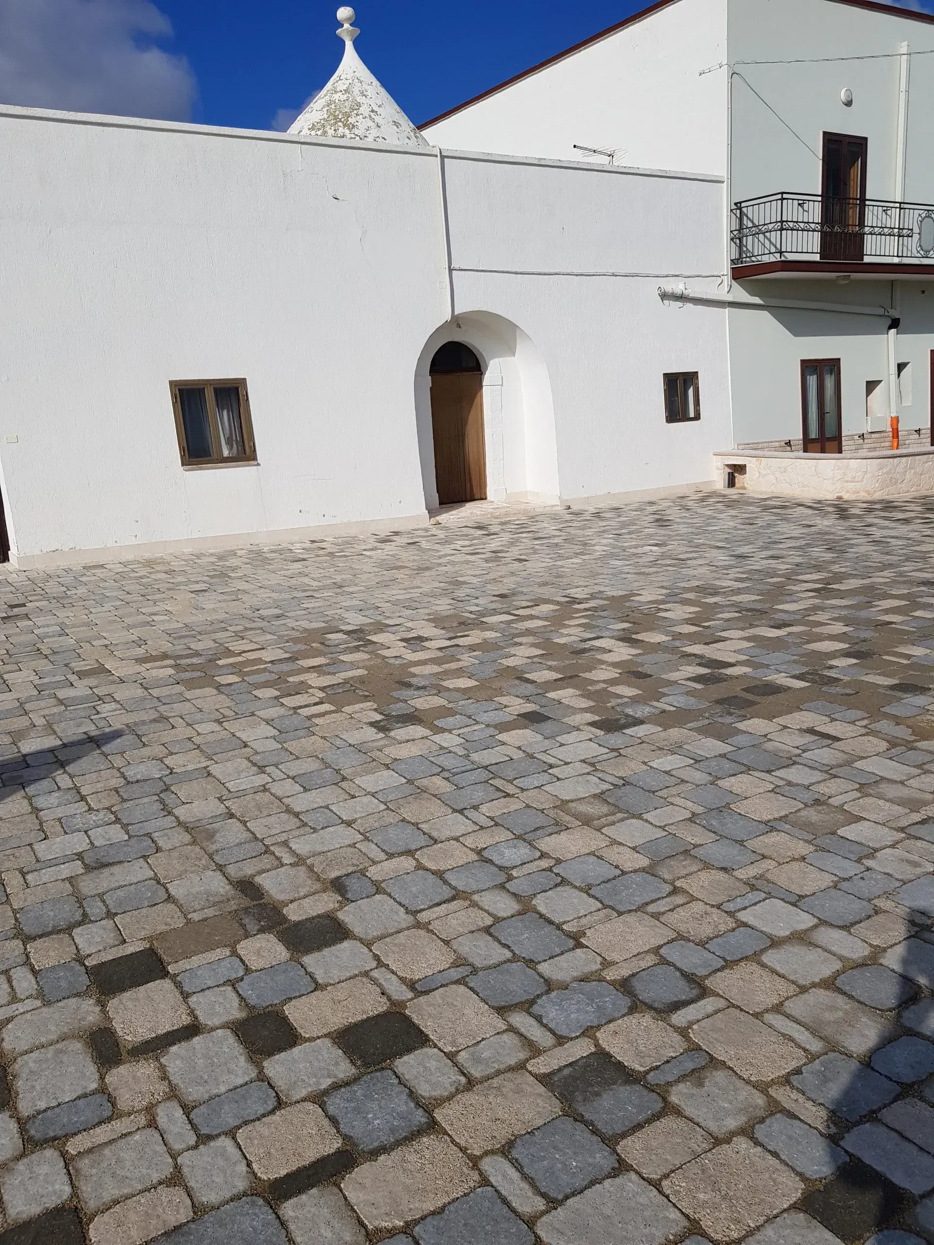 Cortile acciottolato con edifici bianchi e porta ad arco sotto il cielo azzurro.