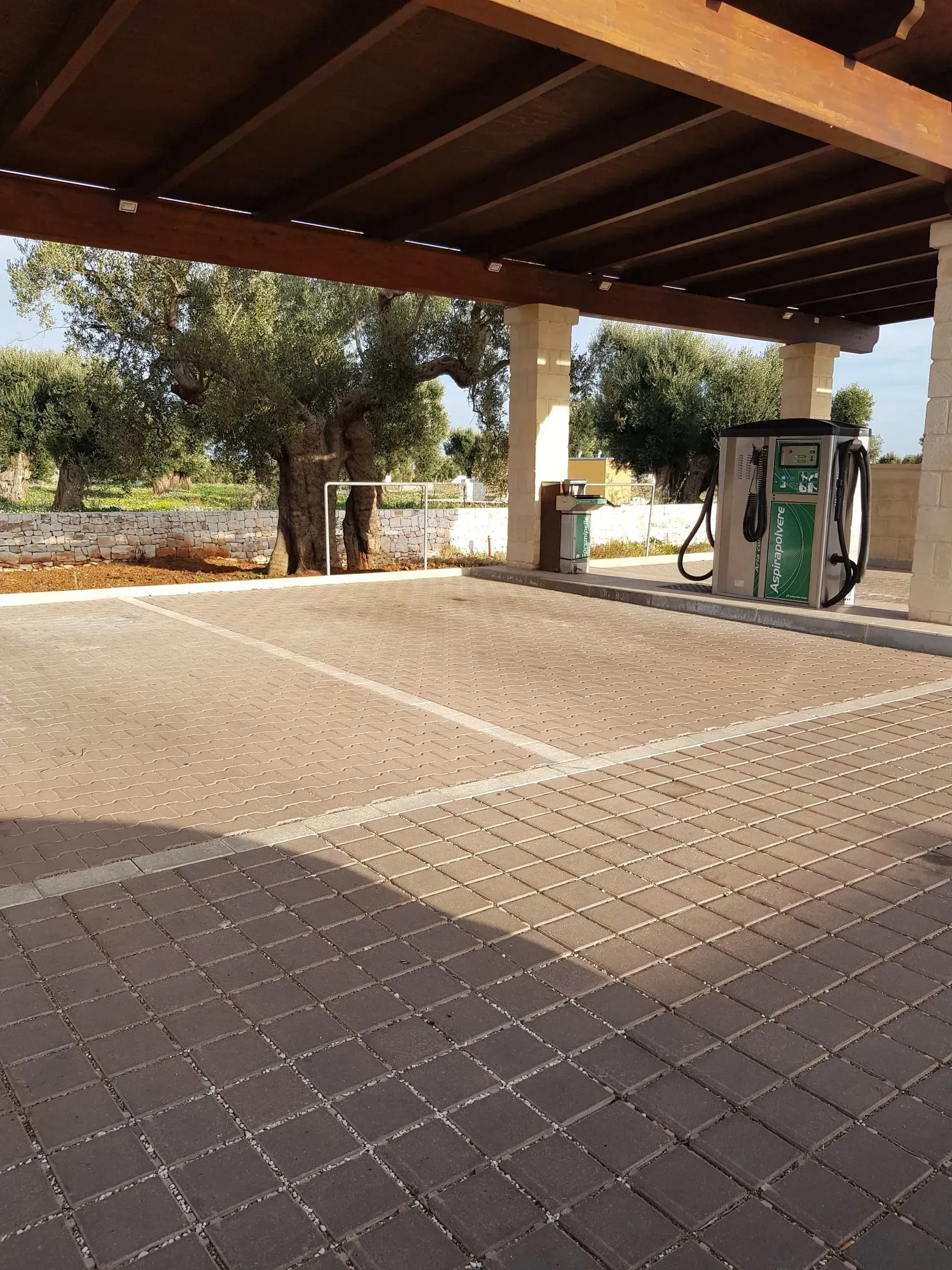Parcheggio coperto con pavimentazione in mattoni, stazione di ricarica e ulivo sullo sfondo.