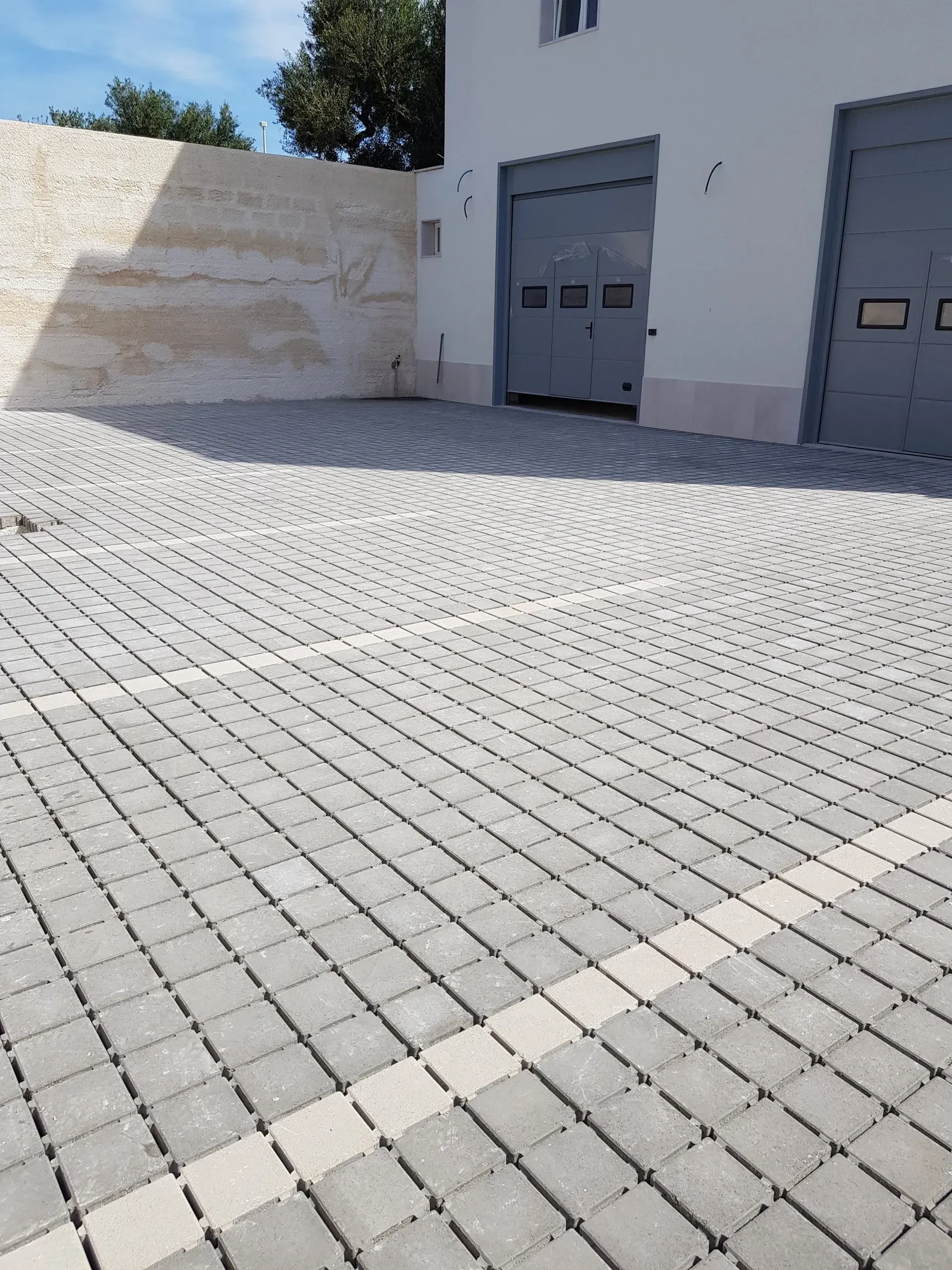 Vialetto in mattoni che conduce alle porte del garage, muro chiaro sulla sinistra, cielo azzurro sopra.