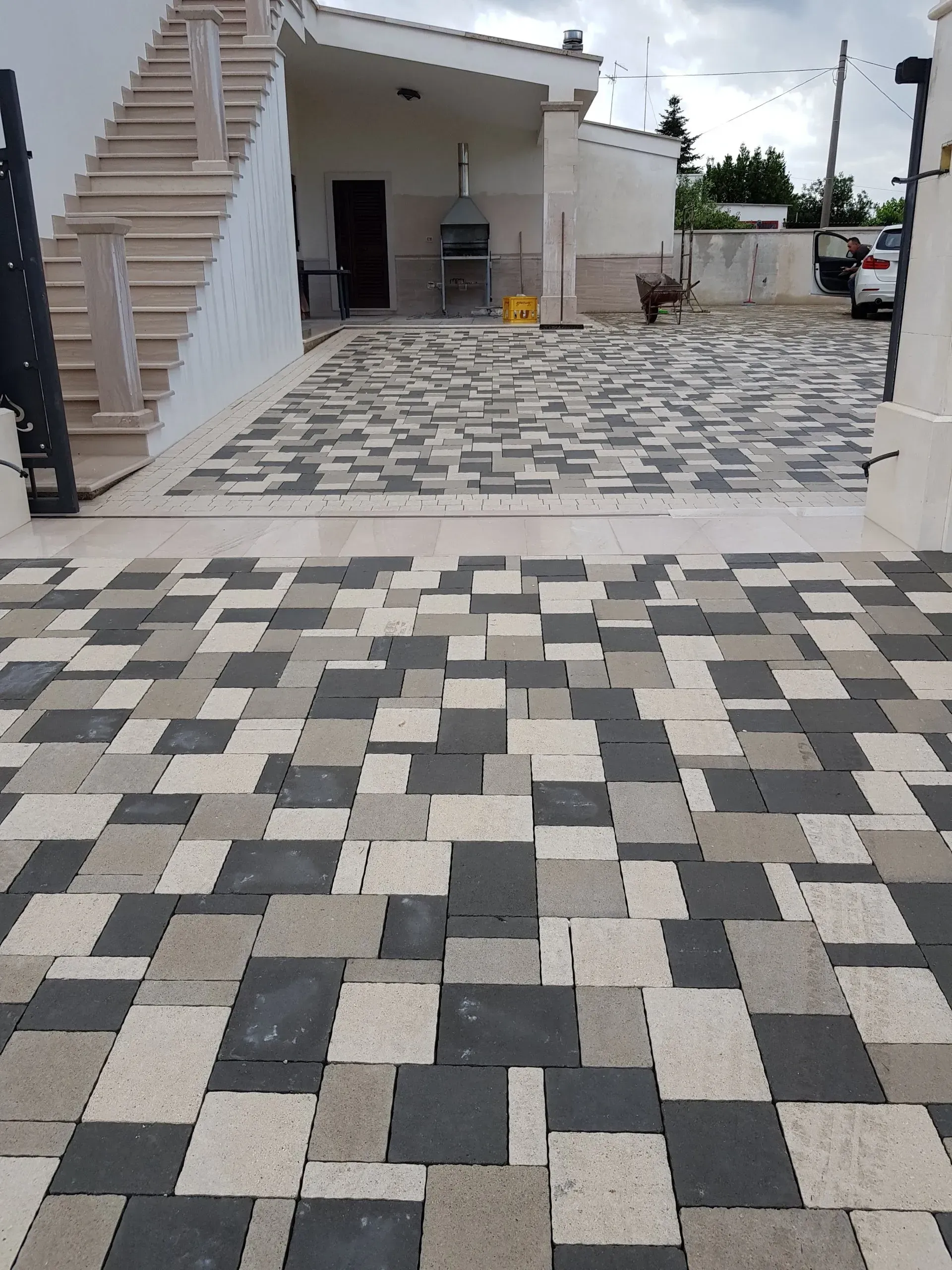 Vialetto d'accesso pavimentato con piastrelle quadrate alternate nere, grigie e beige, che conduce a una casa con pareti bianche e una scala.