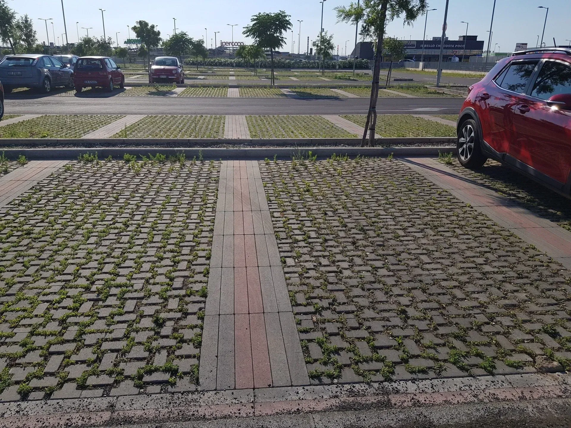Parcheggio pavimentato con blocchi autobloccanti, alcuni con erba. Auto rossa sulla destra. Auto blu e rosse parcheggiate sullo sfondo.