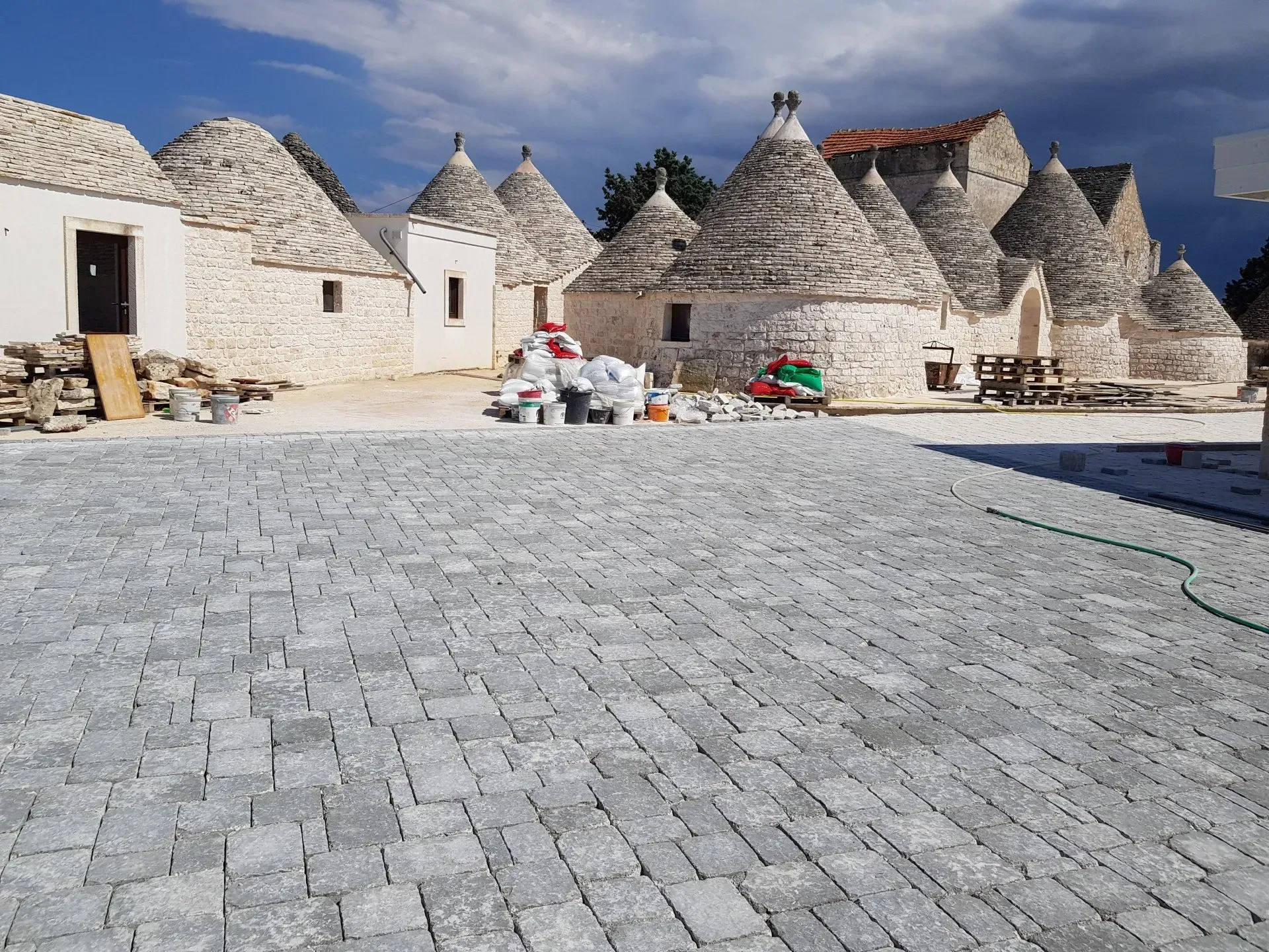 Cortile lastricato in pietra con tradizionali case a forma di cono (trulli) ad Alberobello, Italia, sotto un cielo parzialmente nuvoloso.