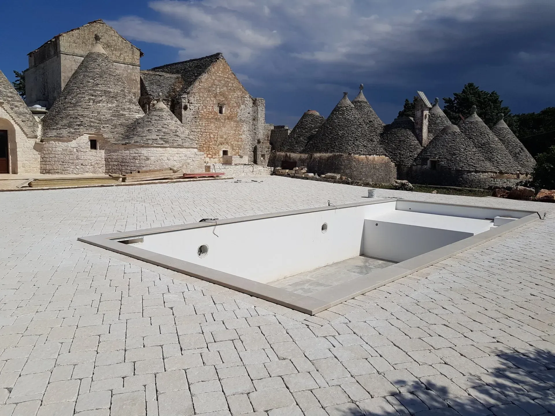 Una piscina rettangolare circondata da lastricati in pietra bianca, con sullo sfondo i tradizionali edifici con tetto conico.
