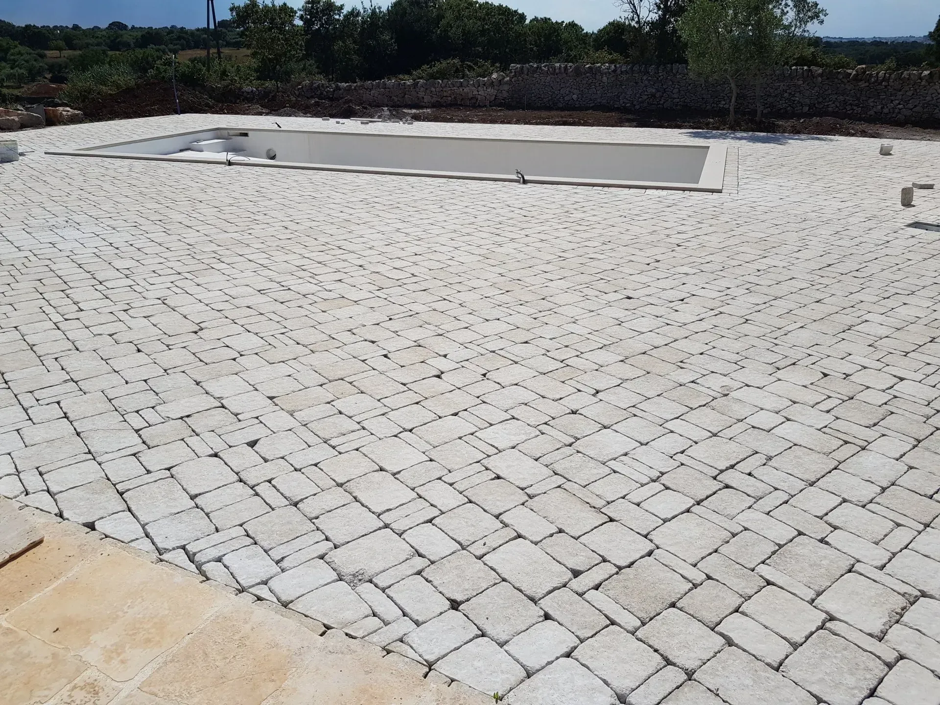 Patio pavimentato in pietra attorno a una piscina rettangolare. Pietre chiare ricoprono il terreno, alberi e cespugli sullo sfondo.