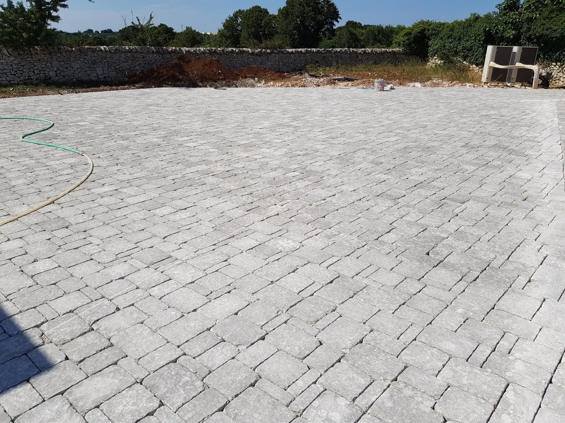 Patio pavimentato in grigio, probabilmente adibito a parcheggio, con un muro in pietra e alberi sullo sfondo.