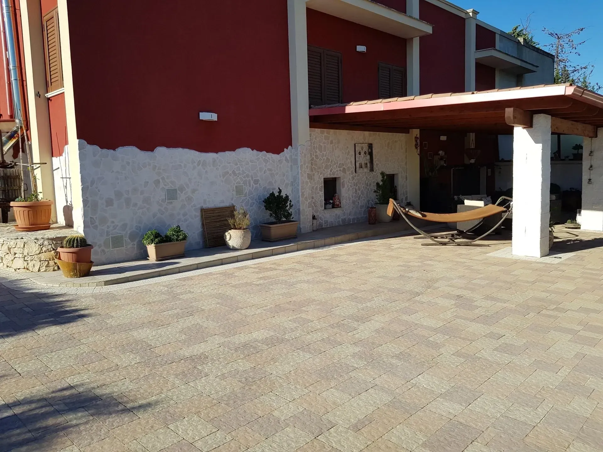 Edificio rosso e bianco con patio coperto, amaca e piante in vaso su un vialetto lastricato in pietra.