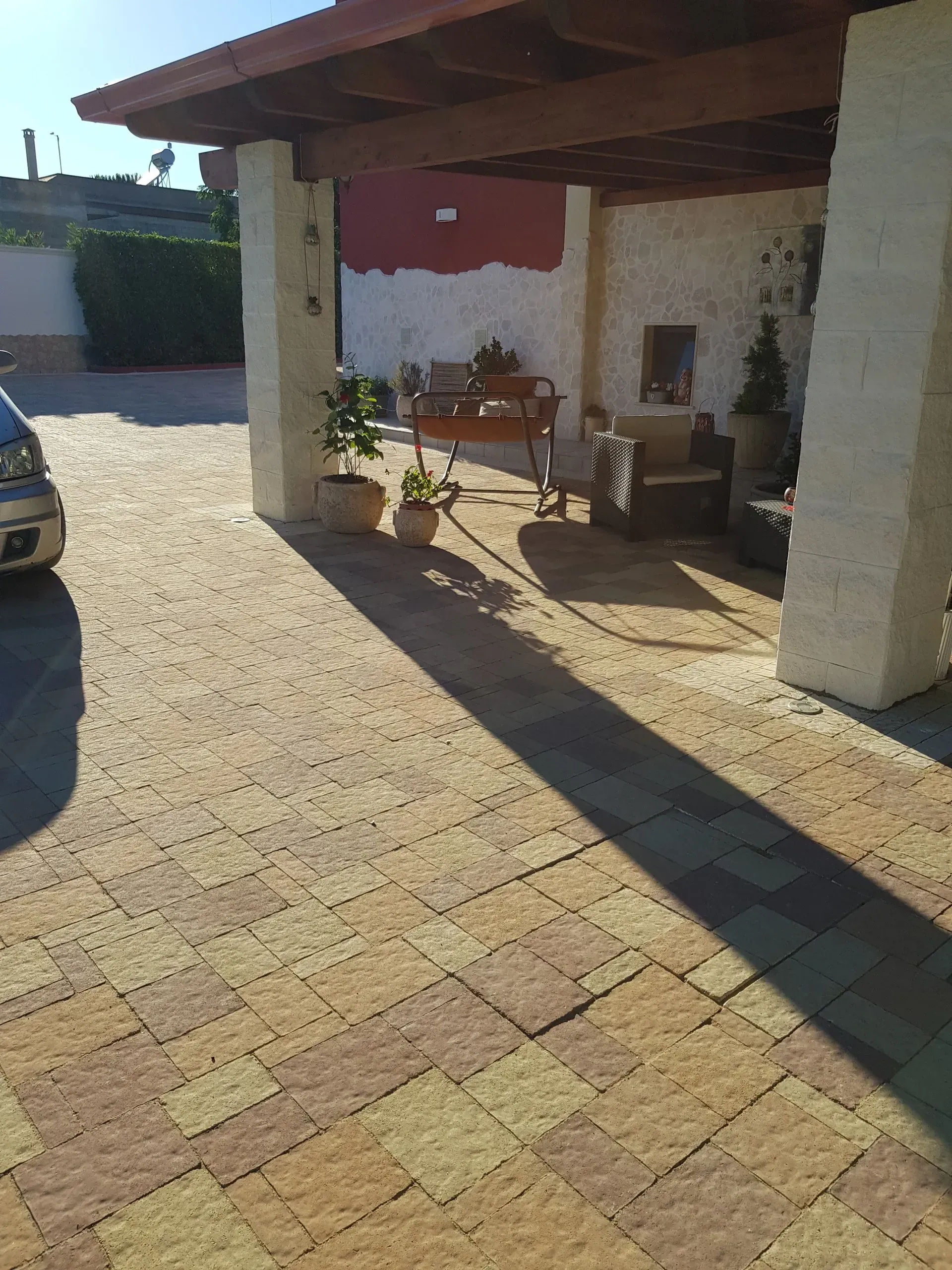 Vista esterna di un patio con pavimentazione in pietra, pilastri e una zona salotto ombreggiata. Un'auto è parcheggiata sulla sinistra.