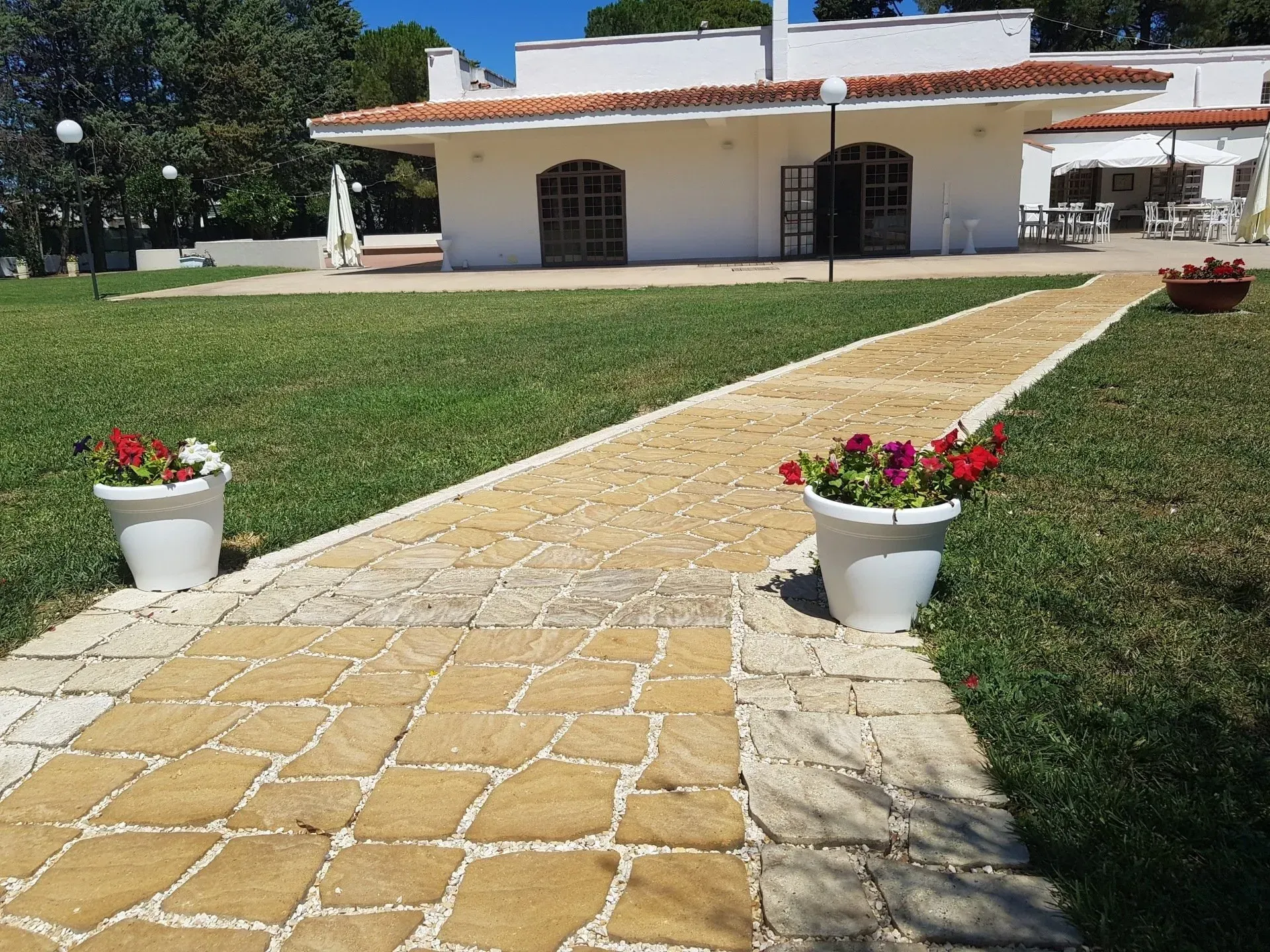 Sentiero in pietra che conduce a un edificio bianco con fiori rossi in vasi disposti accanto all'erba verde.
