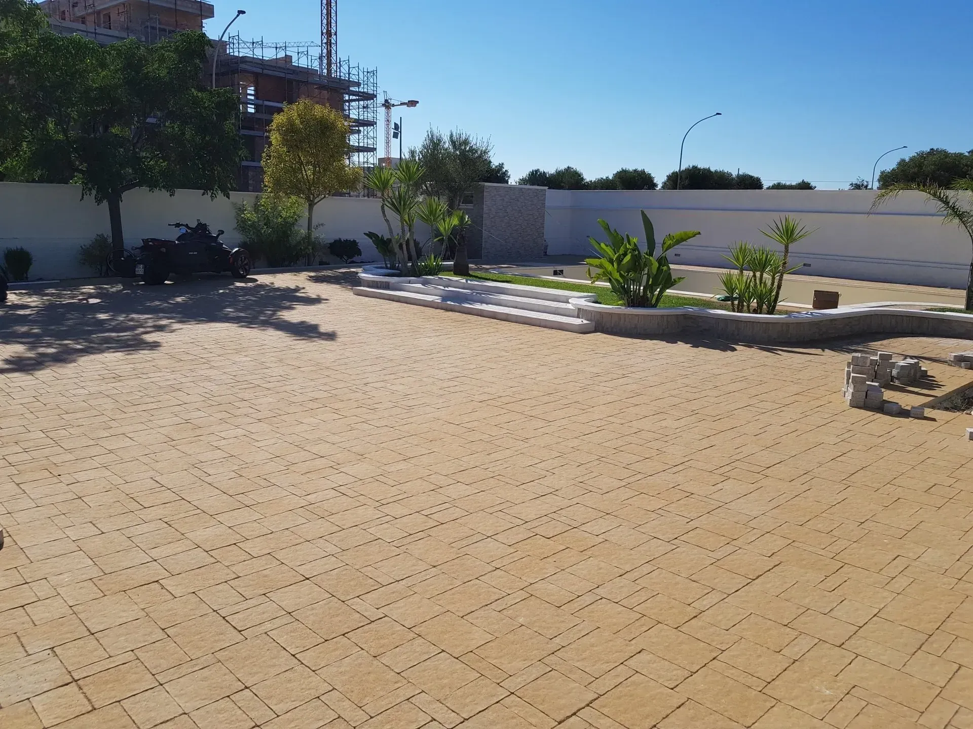 Patio pavimentato in mattoni color cuoio con piccole aiuole, muro bianco e alberi radi sotto un cielo azzurro.