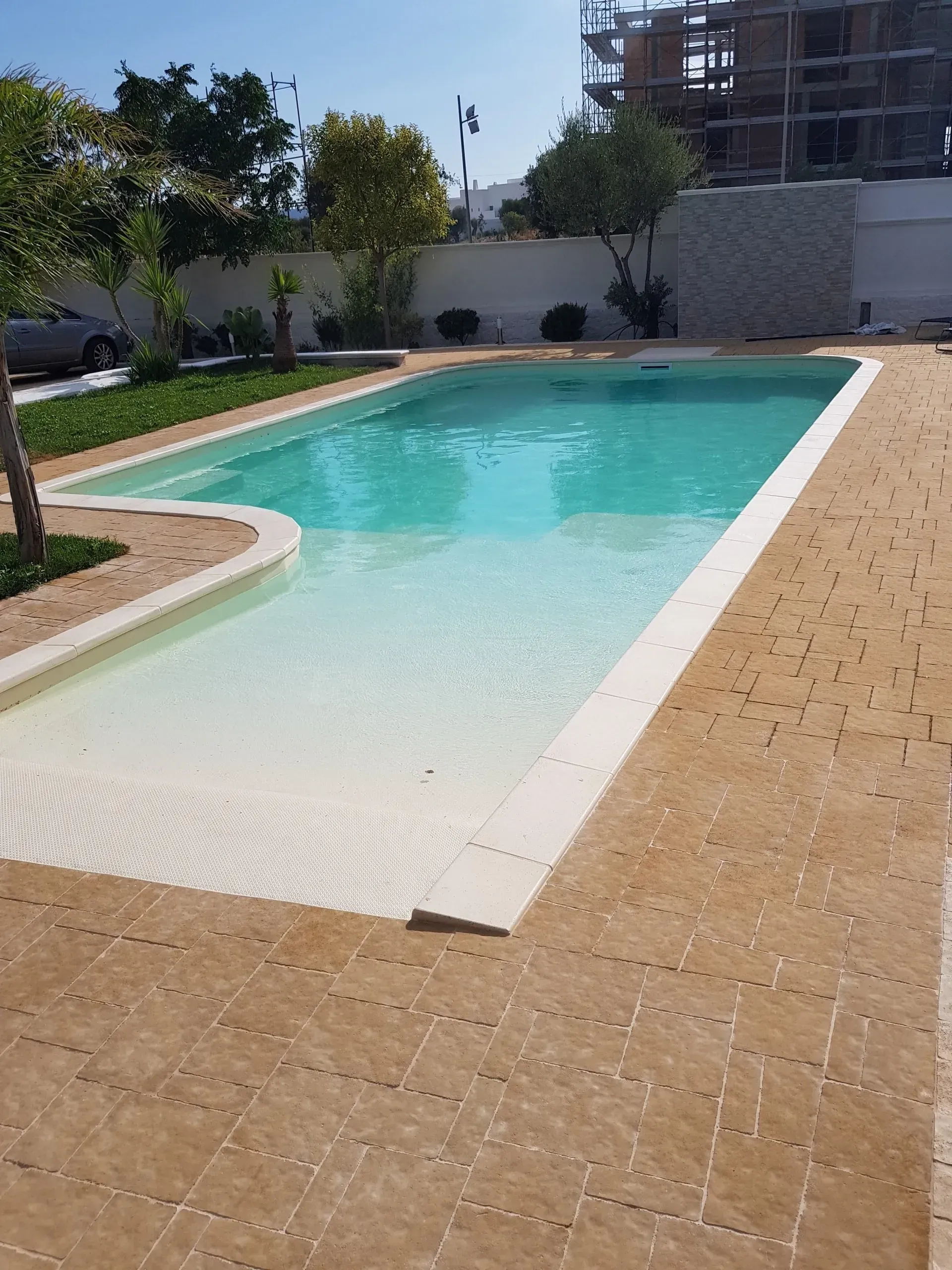 Una piscina rettangolare con ingresso poco profondo, circondata da lastricati beige, in una giornata di sole.