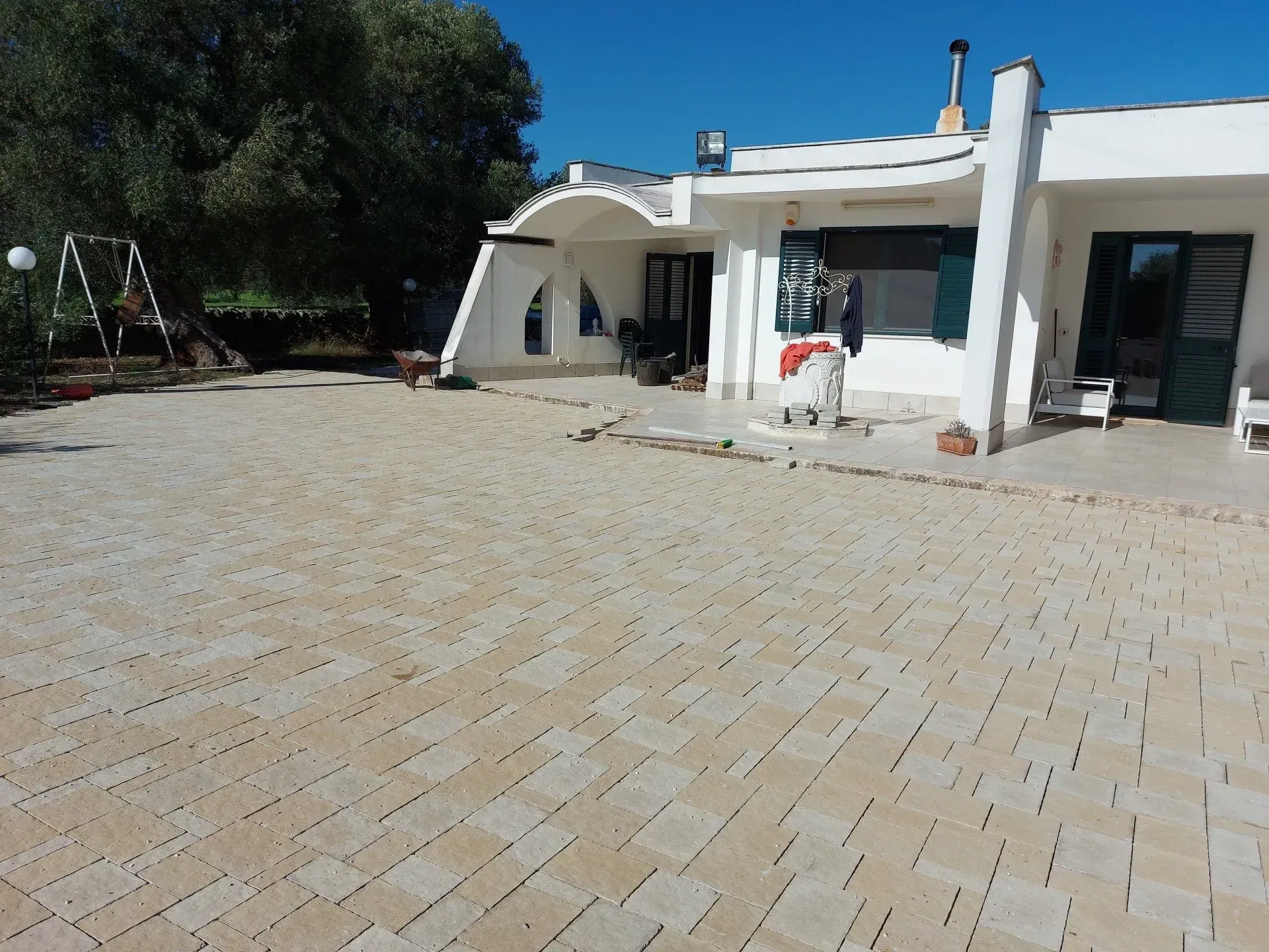 Patio in mattoni beige di fronte a una casa bianca con tetto curvo. Si vedono persiane verdi e un'altalena.