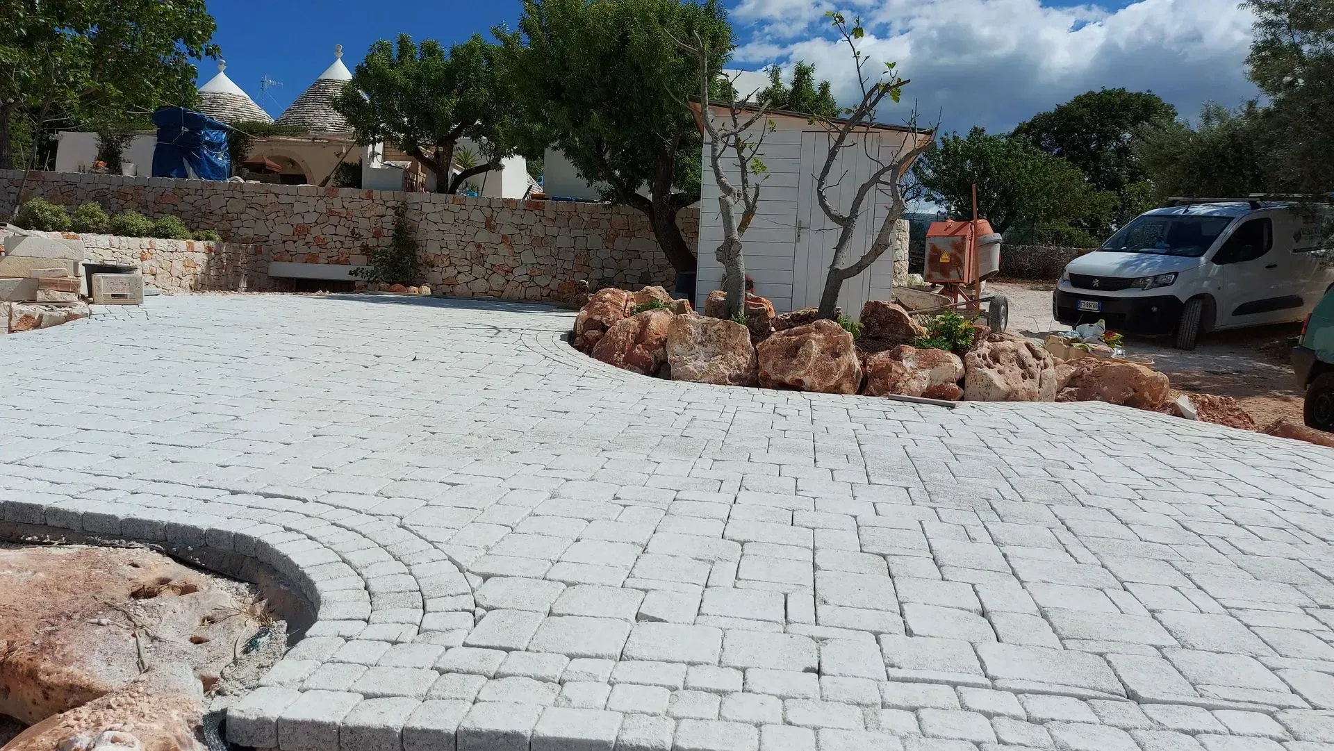 Un vialetto di mattoni bianchi curva verso un edificio, con vegetazione e un veicolo sullo sfondo.