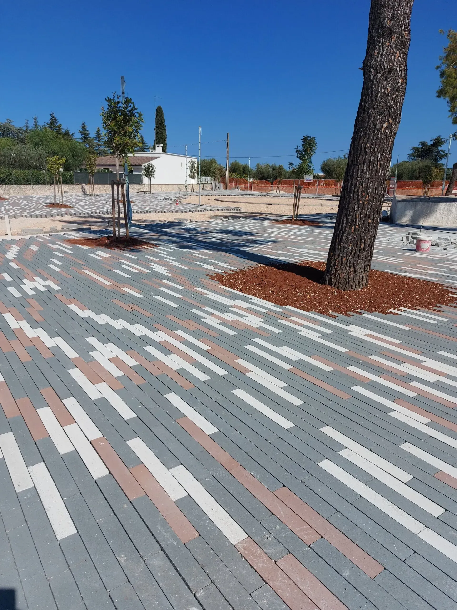 Piazza lastricata di mattoni con alberi e cielo azzurro. Mattoni grigi, bianchi e rossi.