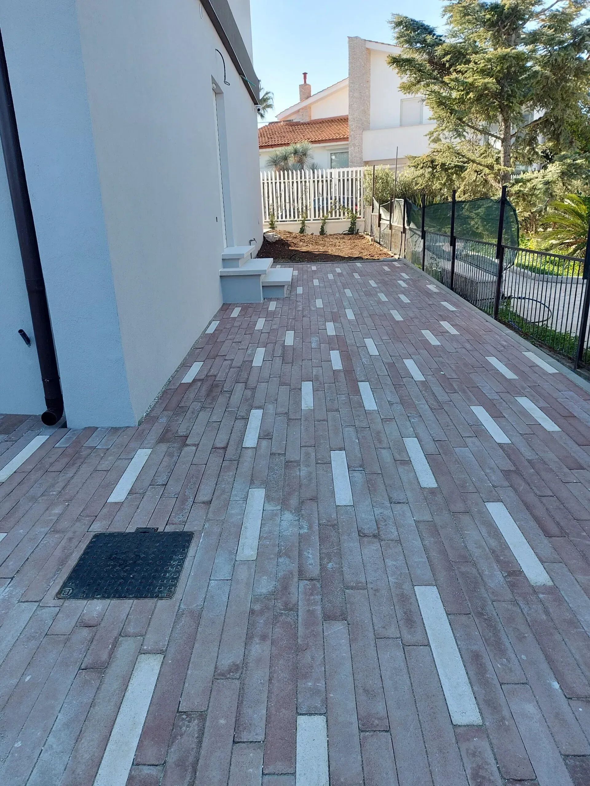 Sentiero in mattoni lungo una casa chiara. Il sentiero è caratterizzato da un motivo in mattoni rossi con accenti bianchi.