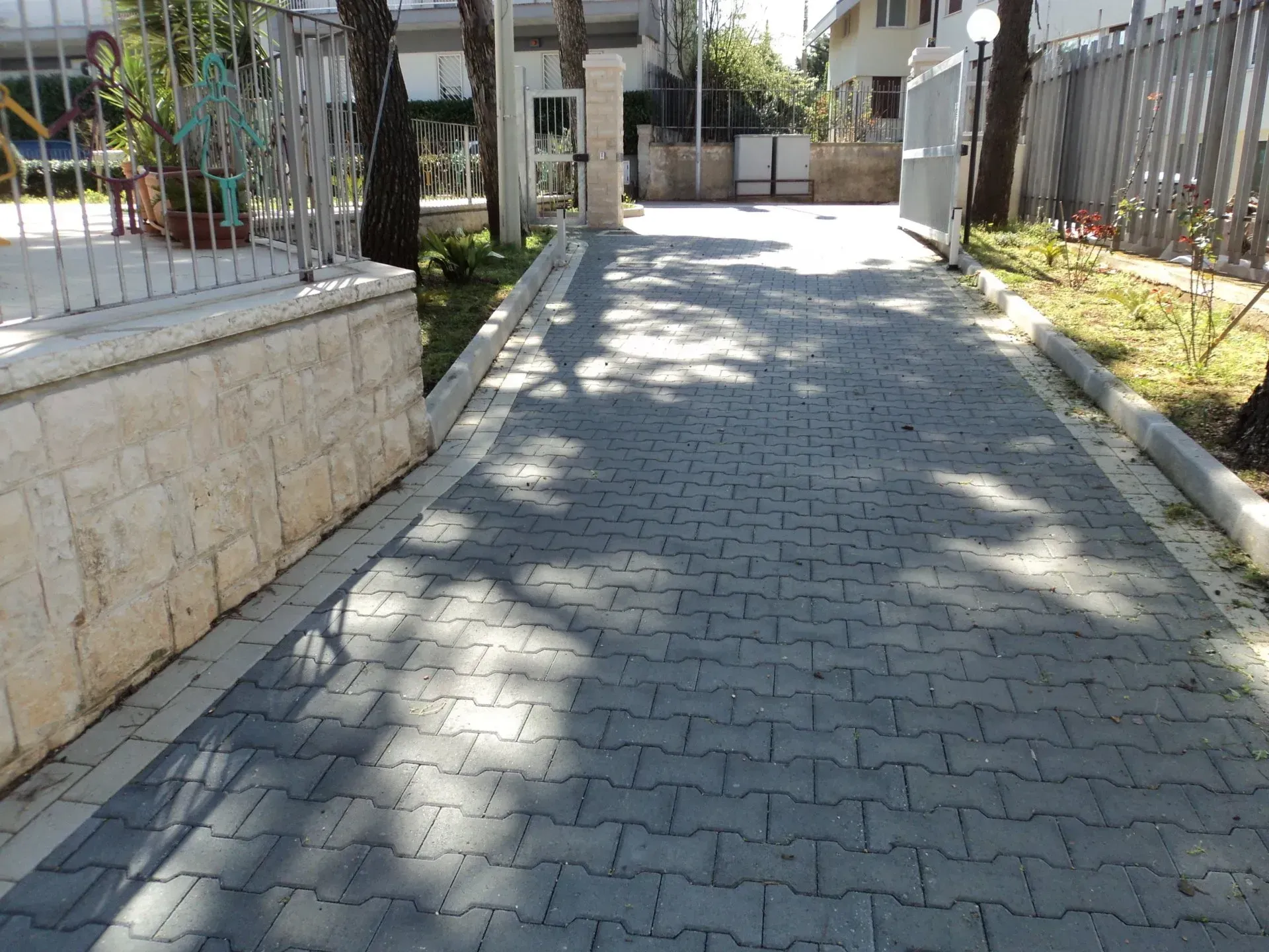 Vialetto d'accesso pavimentato con mattoni grigi ad incastro, delimitato da cemento e da una recinzione, con alberi che proiettano ombre.