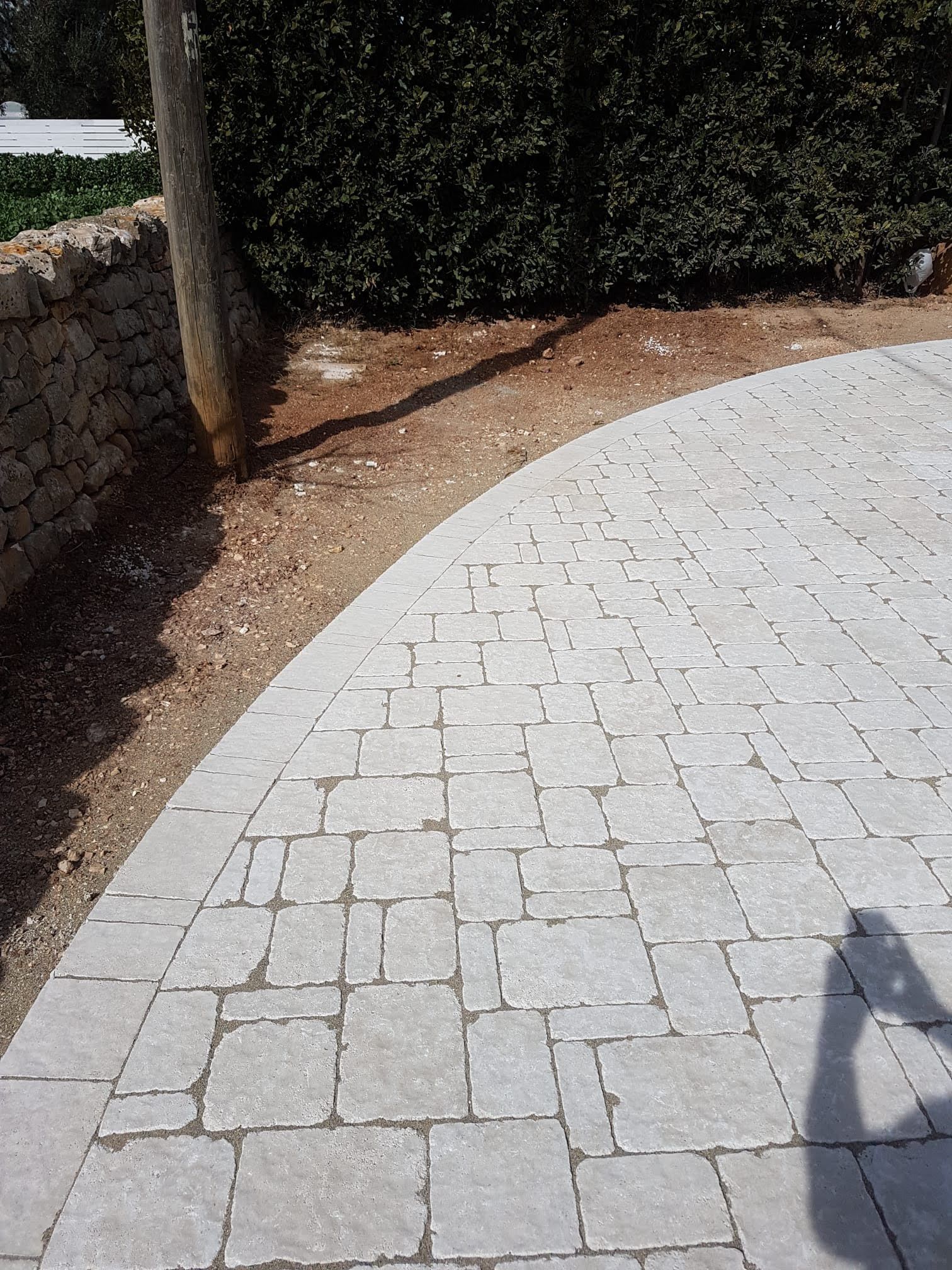 Sentiero curvo in pietra accanto a un bordo sterrato, un muro in pietra e una siepe; la luce del sole crea ombre.