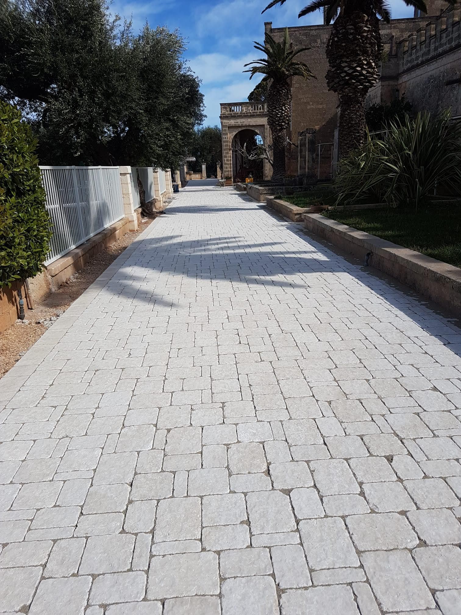 Sentiero in pietra che conduce a un cancello ad arco, fiancheggiato da vegetazione ed edifici sotto un cielo soleggiato.