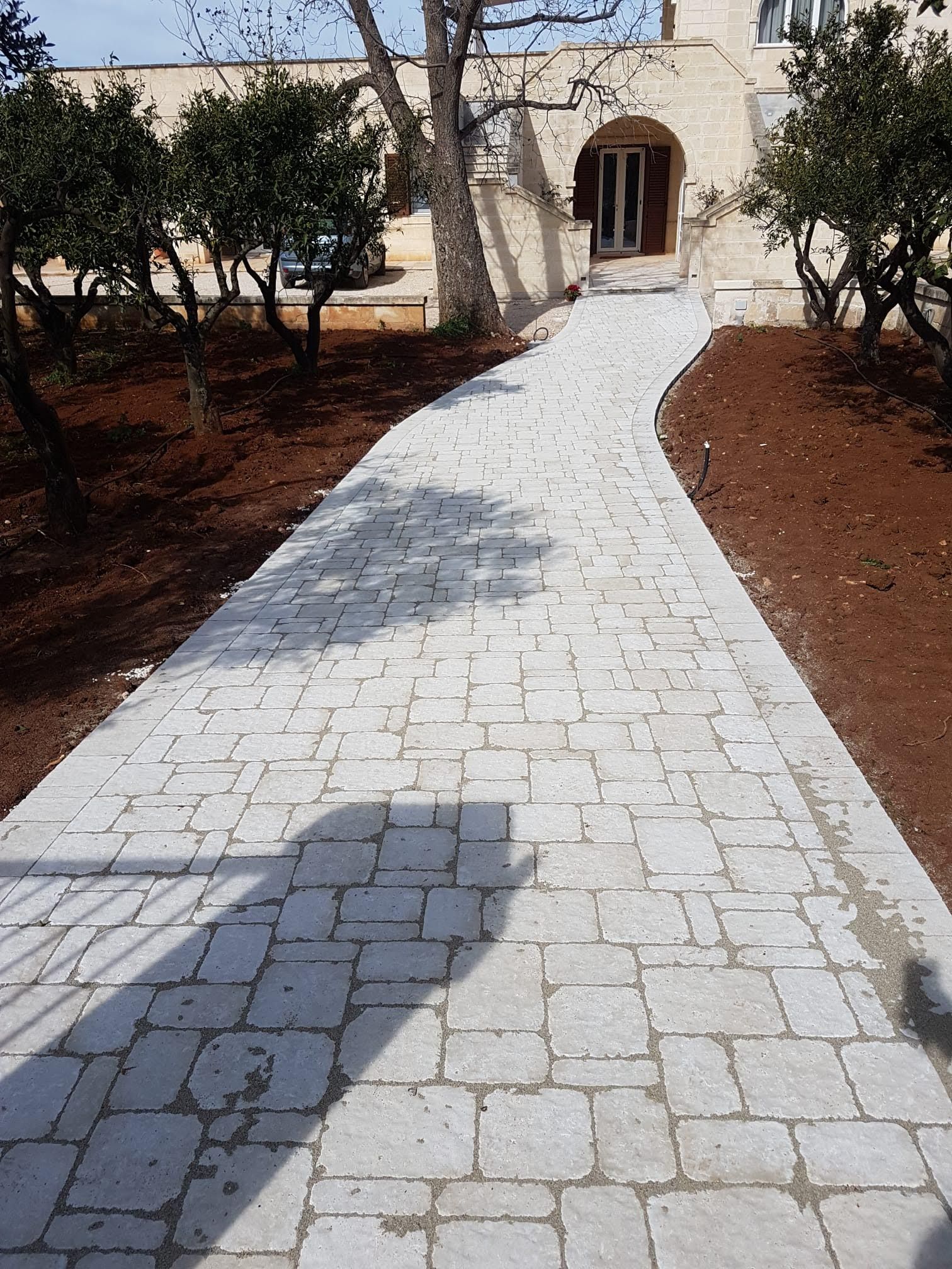 Un sentiero in pietra curva verso un edificio con un ingresso ad arco; alberi e terra scura lo delimitano.