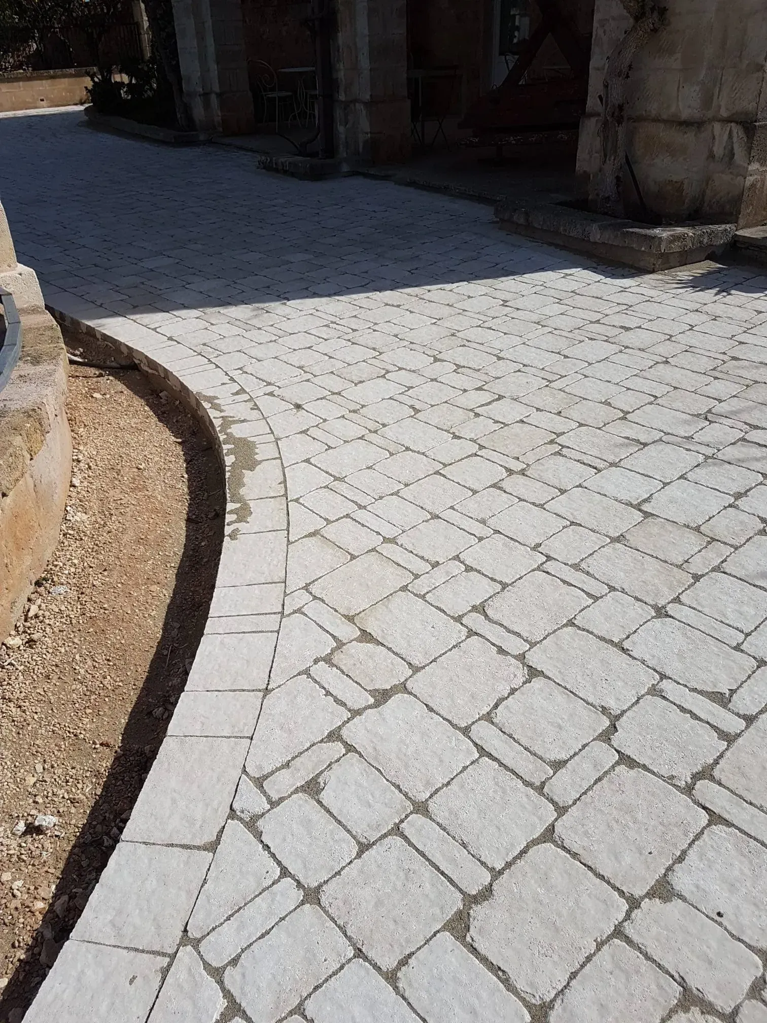 Passerella pavimentata in pietra grigio chiaro con bordo curvo, adiacente a un tratto di terra e a un edificio.