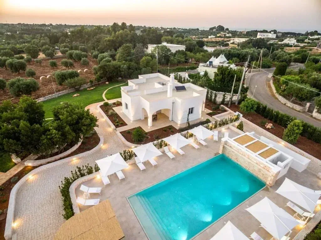 Edificio bianco con piscina e sedie a sdraio; ulivi e strada sullo sfondo.