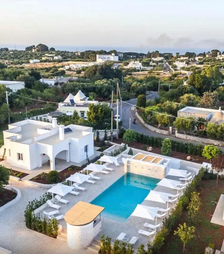 Vista aerea di un resort bianco con piscina, sedie a sdraio ed edifici immersi in un paesaggio rurale verdeggiante.