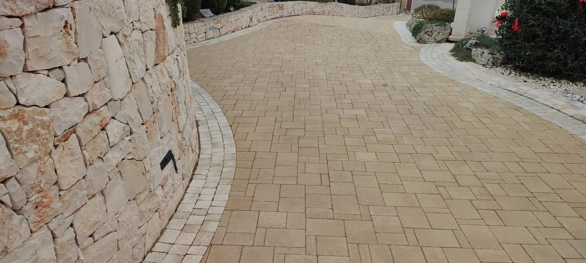 Muro di pietra che si snoda lungo un sentiero lastricato con bordura da giardino e arbusti.