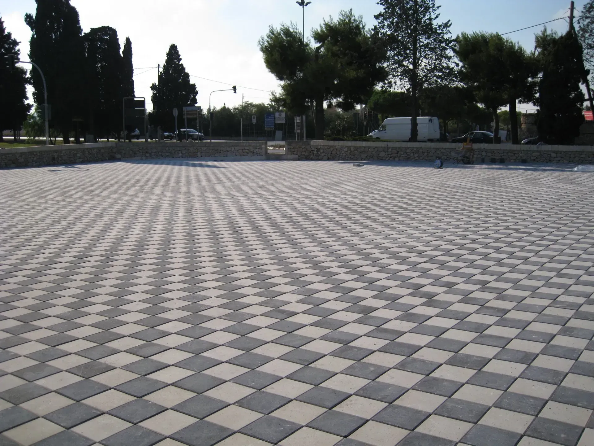 Pavimentazione a scacchiera in uno spazio pubblico, con alberi ed edifici sullo sfondo.