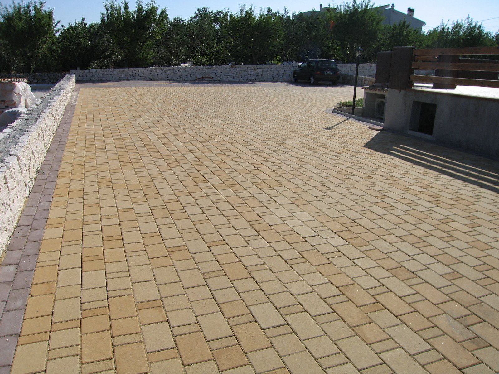 Vialetto lastricato in mattoni, color marrone chiaro, che conduce a una piccola auto parcheggiata, con alberi sullo sfondo.