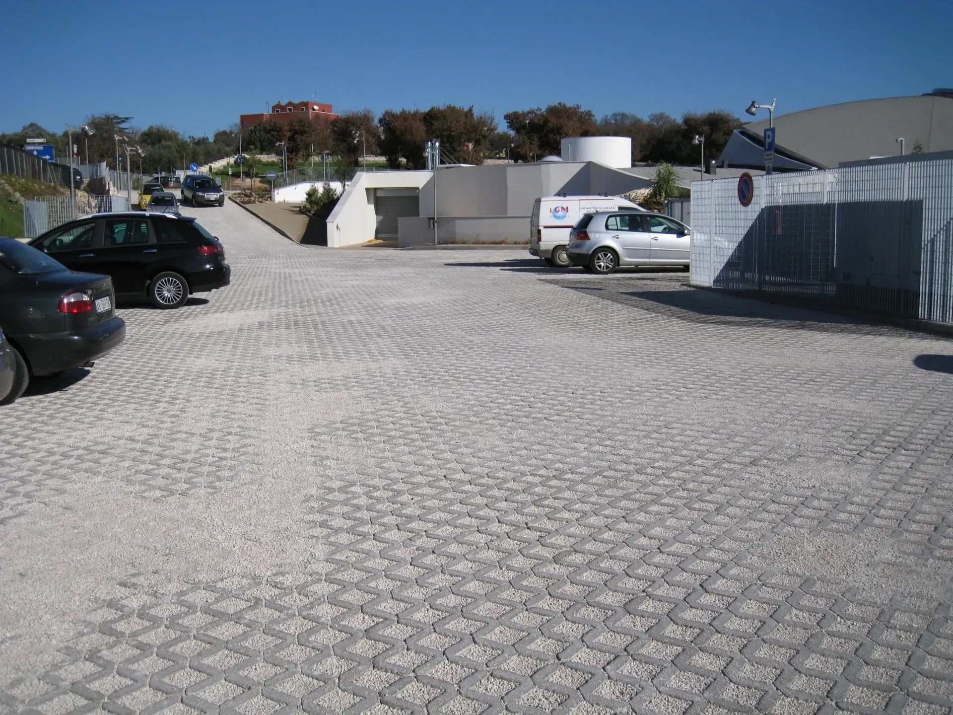 Parcheggio pavimentato con blocchi di cemento autobloccanti; auto parcheggiate; edifici sullo sfondo.
