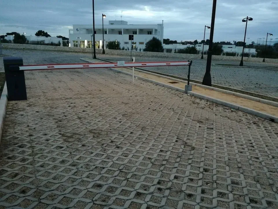 Ingresso del parcheggio con barriera, pavimentazione in cemento e un edificio bianco sullo sfondo.