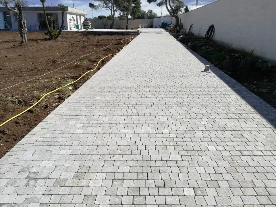 Sentiero in blocchi di pietra che conduce a un edificio bianco con alberi in un ambiente esterno soleggiato.