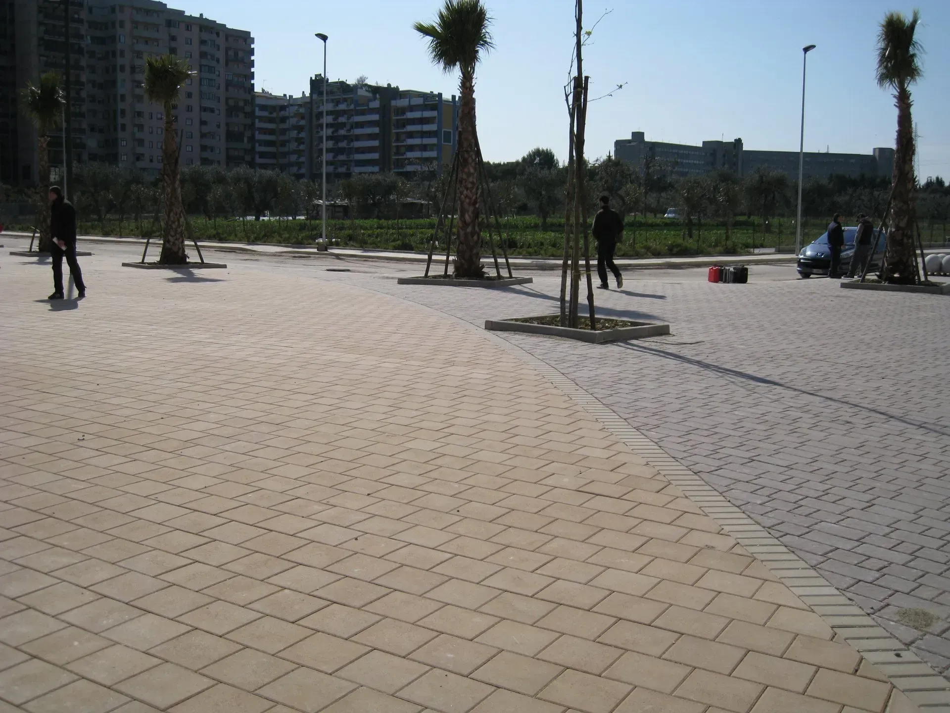 Area pavimentata con alberi ed edifici sullo sfondo; due persone che camminano, giornata di sole.