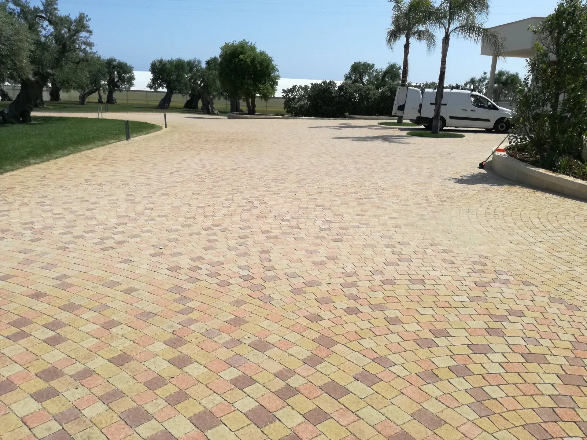 Parcheggio pavimentato in mattoni con un furgone bianco, alberi ed erba sotto un cielo luminoso.