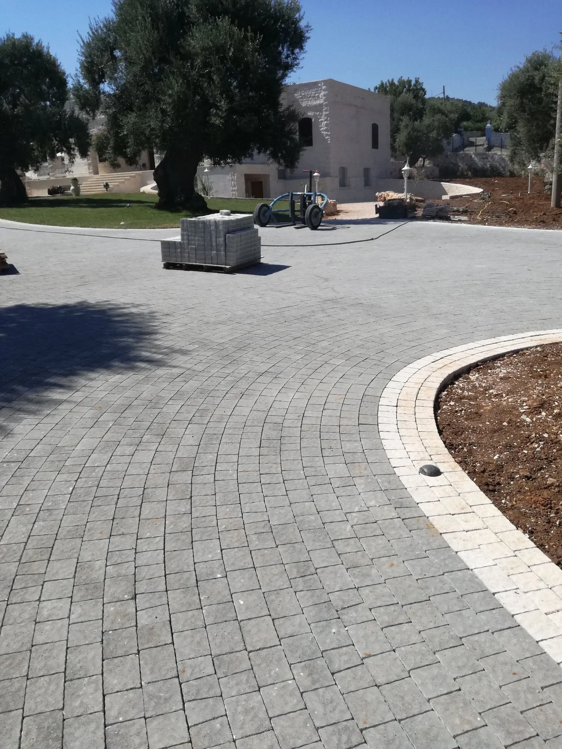 Area pavimentata in ciottoli grigi con bordo curvo, attrezzature edili e un edificio bianco sullo sfondo.